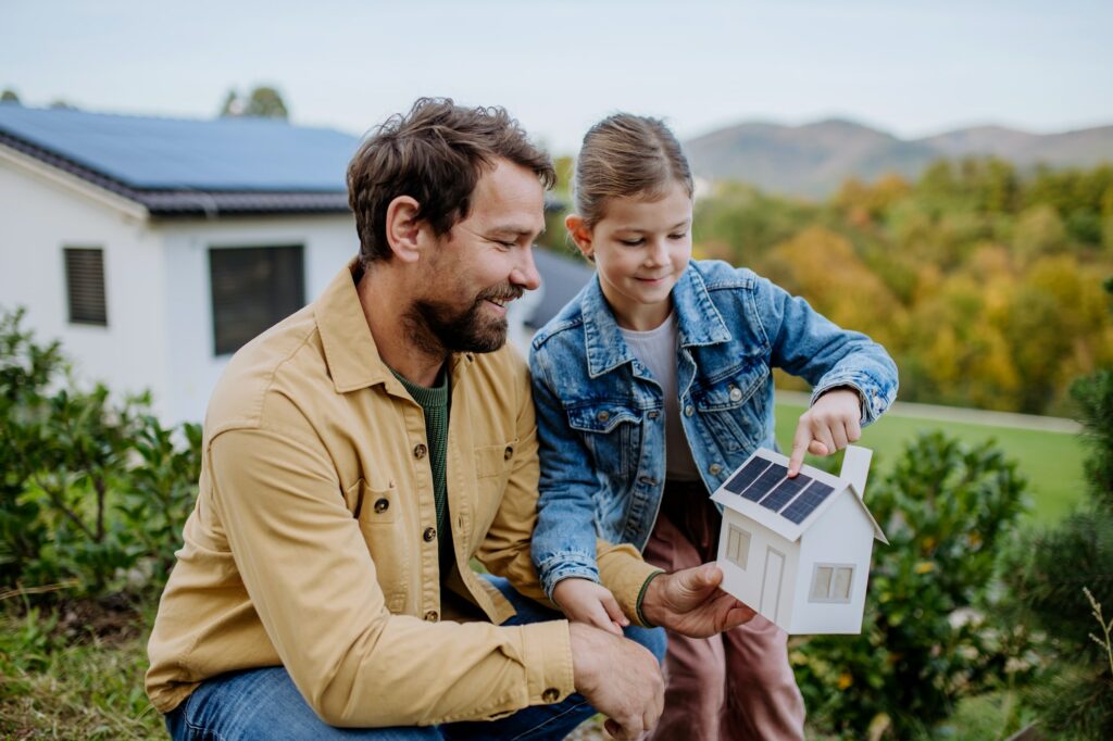 Kleines Mädchen mit ihrem Vater, der ein Papiermodell eines Hauses mit Sonnenkollektoren hält und erklärt, wie es funktioniert