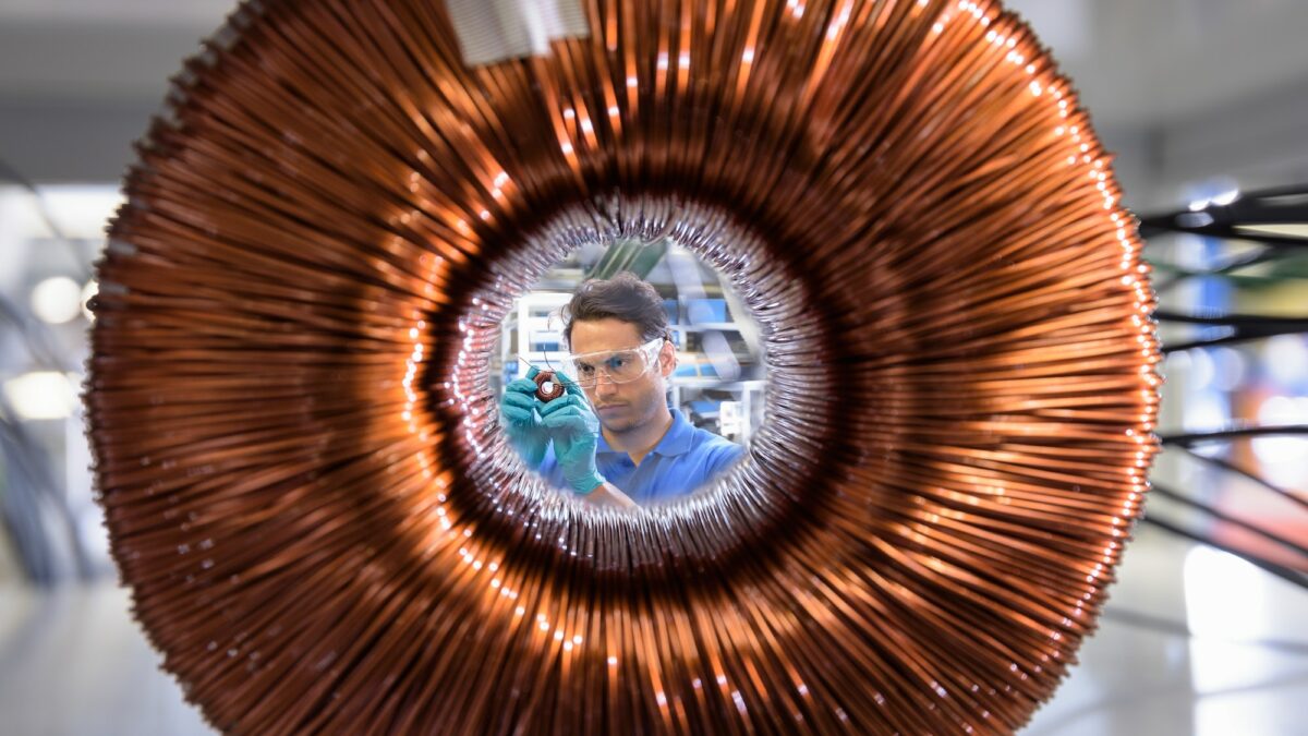 Worker inspecting electromagnetic coil seen through large coil in electromagnetics factory