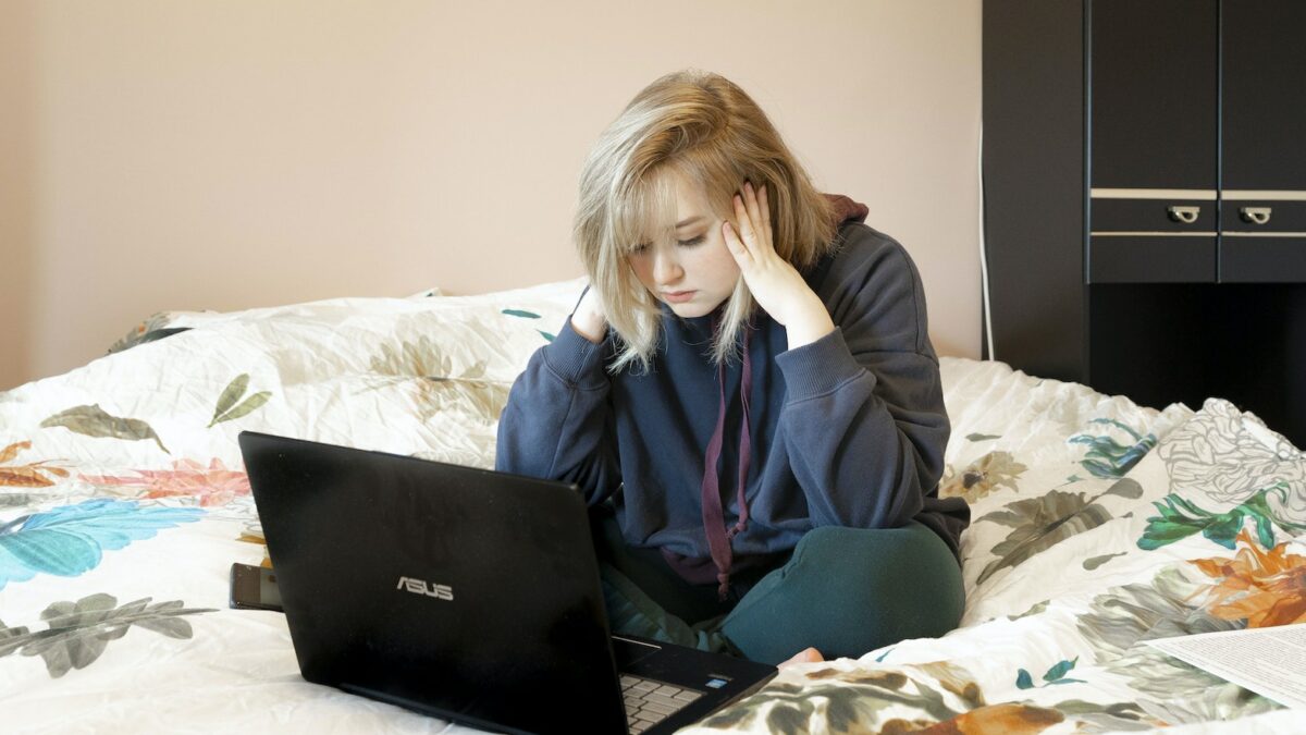 Woman in Blue Hoodie Sitting on Bed Using Black Laptop Computer