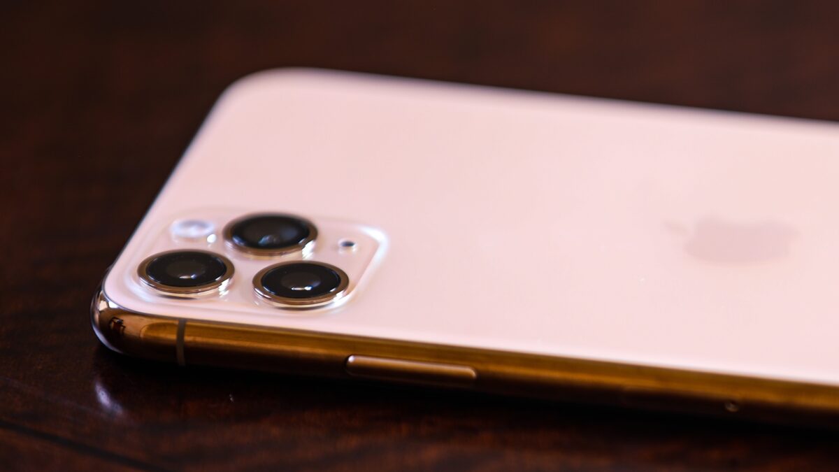 iPhone11 Pro Max in gold with focus on the triple lens. Placed on a mahogany wood table.