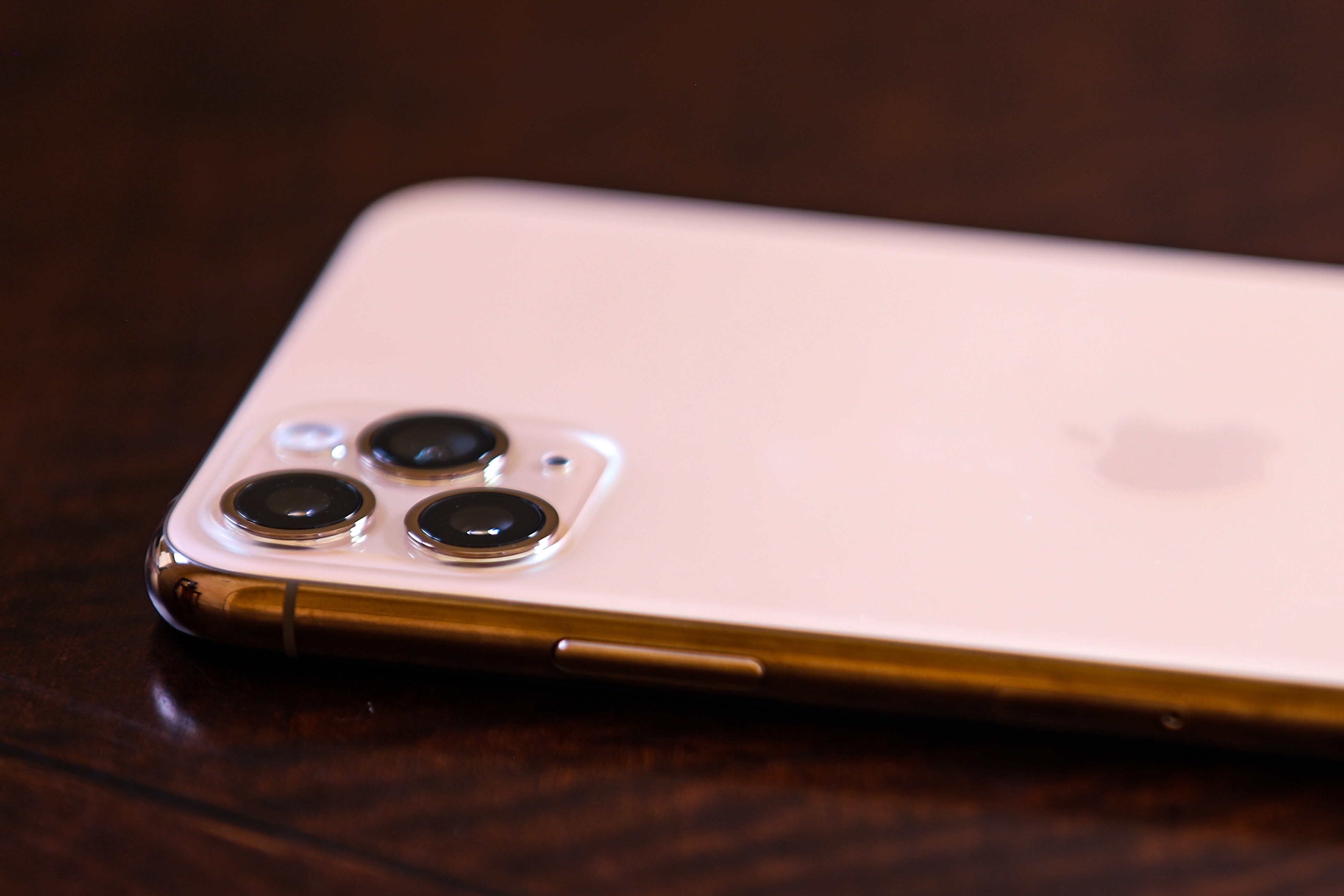 iPhone11 Pro Max in gold with focus on the triple lens. Placed on a mahogany wood table.